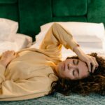 Woman lying on bed with hand on forehead, appearing relaxed or sleepy in a cozy indoor setting.