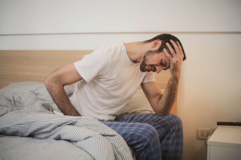 Side view of male in sleepwear sitting on bed with closed eyes after awakening and suffering from migraine while touching head and grimacing in pain